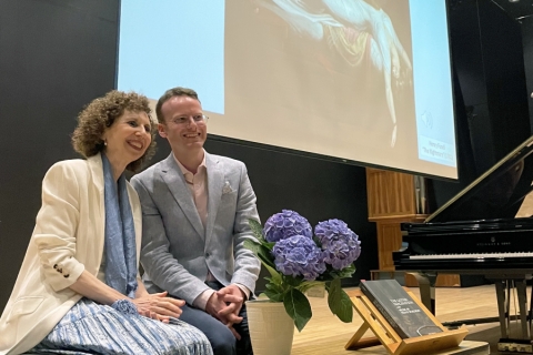 Two people on stage next to purple flowers.